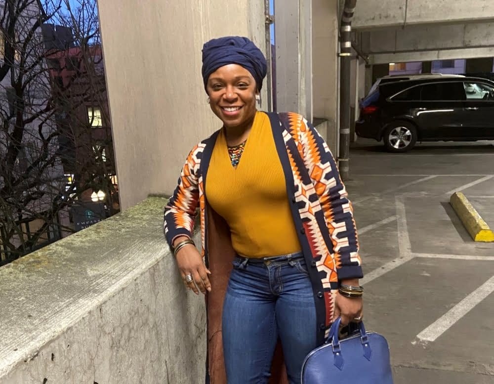 Healthcare accountant Lozelle Mathai wearing yellow v-neck shirt and jeans with patterned cardigan, head wrap, and blue bag in parking garage with visibile trees and black car at dusk